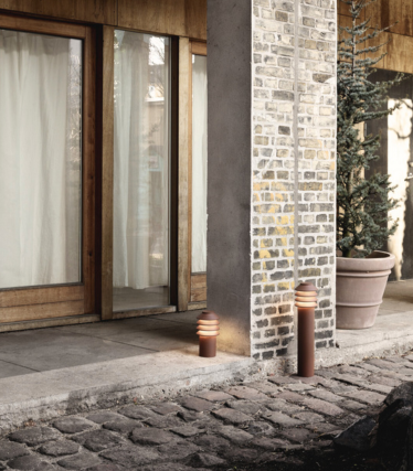 Exteriérová scéna s dvomi modernými valcovými svetlami pri kamennom chodníku a tehlovo-betónovom stĺpe. V pozadí sú sklenené dvere s dreveným rámom, ľahké závesy a veľká črepníková vždyzelená rastlina. Teplá žiara svetiel vytvára útulnú a príjemnú atmosféru.